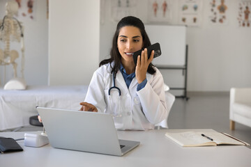 Positive young therapist doctor woman speaking to patient on cellphone at workplace, recording...
