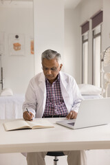 Serious focused older Indian practitioner man in white uniform coat writing medical notes, patients illness history in notebook, working at laptop computer, sitting at workplace table