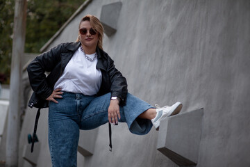 A beautiful plus size woman dressed jeans, jacket, white t-shirt, accessories. Latin american girl walking against grey wall on the city street
