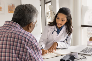 Fototapeta premium Serious young doctor woman writing down patients complaints, listening to older man visiting practitioner office, giving medical consultation, recommendation for checkup, treatment
