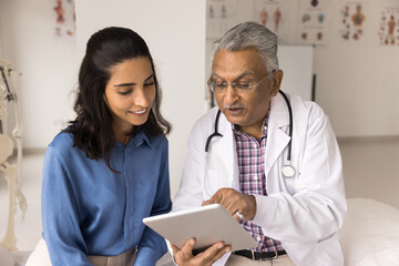Positive grey haired Indian practitioner doctor man showing medical test results on tablet to young patient woman, speaking, explaining electronic content, using digital device for work