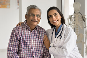 Positive beautiful young medical professional woman embracing elderly Indian patient, expressing warmth, care, support, empathy, looking at camera with toothy smile, posing for portrait