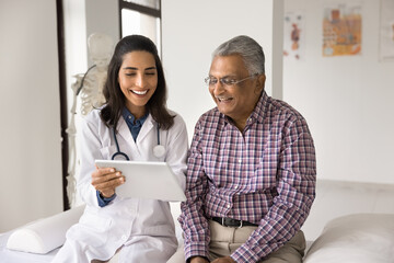 Happy geriatric young doctor and senior Indian patient man discussing modern technology, healthcare, using digital tablet together, looking at display in practitioner office, laughing - obrazy, fototapety, plakaty