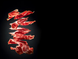 Thin slices of Spanish jamon flying in the air on black background. Traditional meat specialty of the local cuisine - obrazy, fototapety, plakaty