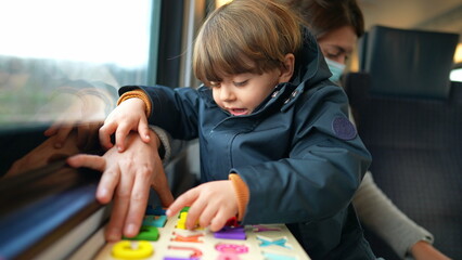 Child seated on mother lap on train ride while doing school lesson activity on the go. Little boy...