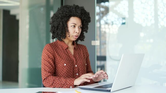 Young african american female employee having a panic attack working on a laptop sitting at workplace in office. Black woman feels fear, breathes hard, is stressed. Shocked lady's fingers are shaking