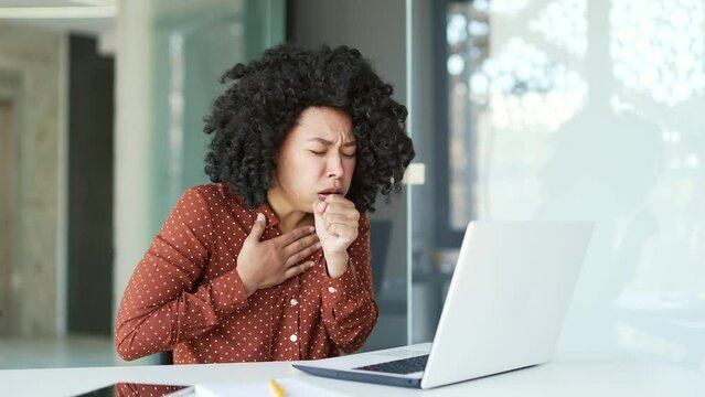 Sick young african american female employee coughing while working on a laptop while sitting at a workplace at desk in office. Tired black woman has the first signs of a cold, virus, bronchitis or flu