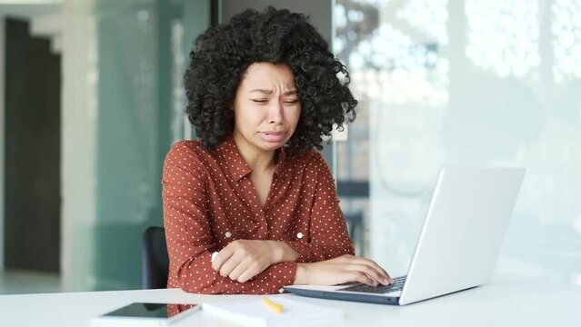 Sad young african american female employee crying and suffering from mental pain sitting at workplace in office. A disappointed black woman has problems at work, worries, feels despair and depression