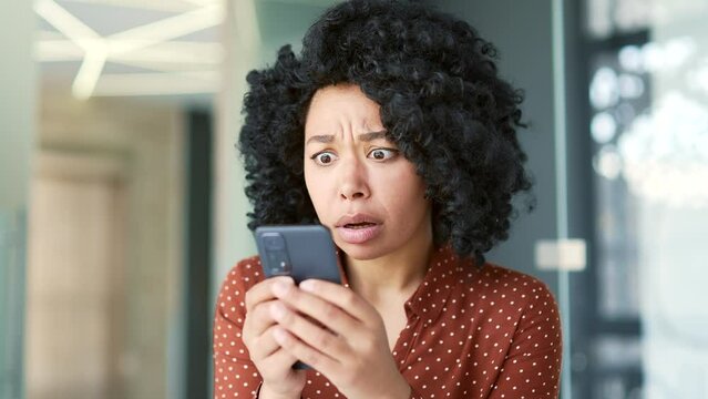 Close up. Upset shocked young african american female employee reads bad news on smartphone at workplace in office. Black woman is disappointed by a message, received notification or seen on Internet