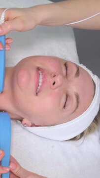 Close-up, a beautician performs microcurrent stimulation on a young woman's face in a spa clinic. Vertical video