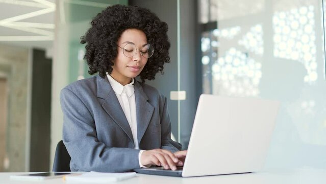 Young african american businesswoman in a suit typing on laptop while sitting at workplace in business office. Black woman in glasses works on computer on project, chats online, writes email messages