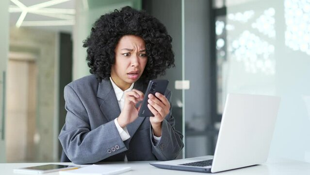 Frustrated young african american businesswoman complains about poor performance of software on smartphone sitting in office. Black woman is dissatisfied with work of phone, the Internet or service