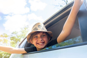emotional boy waves his hands from the car window. happy child in a hat looks out of an open car...