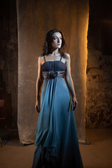 An elegant and beautiful brunette in a chic evening dress, a blue dress with a long flowing skirt to the floor. photo in dark colors on a textured wall, warm light