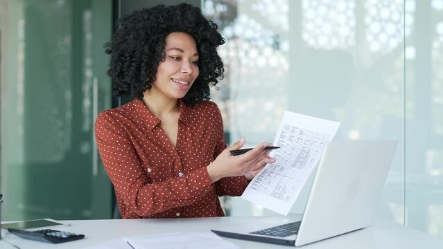 Confident african american female manager talks online via video call using laptop sitting at workplace in office. Financier shows a financial report and statistical documents on a remote presentation
