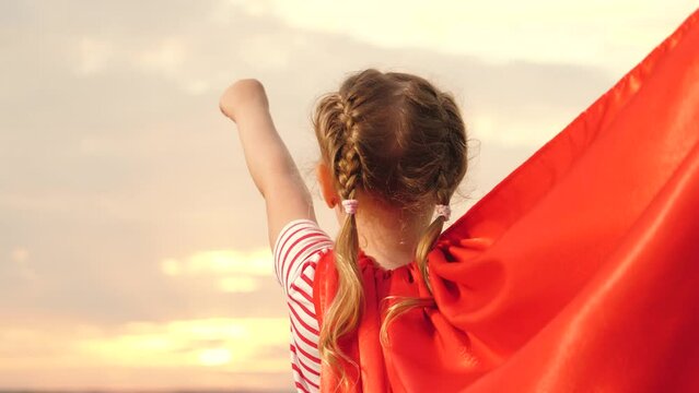 Girl pretends to be superwoman with red cape blowing in wind at sunset on field