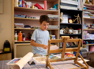 Handsome boy artist standing by desk near a wooden easel, choosing sketch for making image reproduction with watercolors