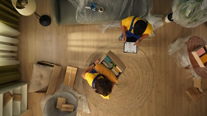 Top view of apartment living room. Delivery service male workers in uniform man holding box with stuff other checking it on list.