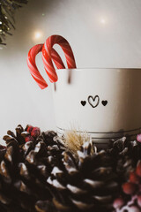 Candy cane sticking out of a cup, warm lights in the background. Pinecones covered in snow,...