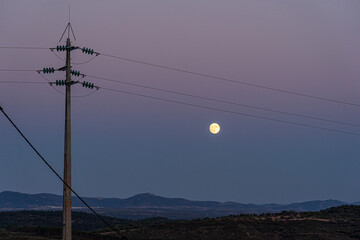 full moon rising behind the mountains at nightfall with high voltage pole in the foreground,...