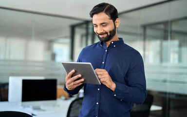 Smiling professional Indian businessman using tab computer working standing in office. Busy...
