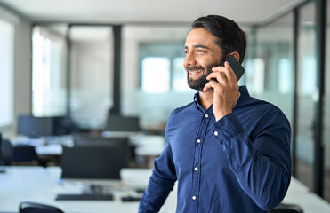 Happy Indian business man talking on mobile cell phone device looking away standing at work. Smiling professional businessman making call on smartphone working with cellphone in office.