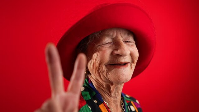 Slow motion of funny, elderly mature woman, 80s, giving peace sign gesture with no teeth and red hat, isolated on red background studio. Concept of youthful old lady