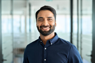 Happy bearded Indian business man leader looking at camera standing in office hallway. Professional smiling businessman manager executive or male employee or entrepreneur headshot portrait.
