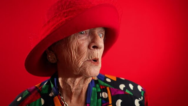 Funny fisheye of elderly senior old woman with wrinkled skin wearing red hat gives great idea gesture showing explosion of thinking posing isolated on red background.