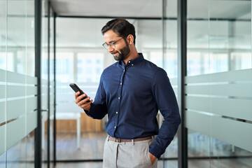 Indian business man employee using mobile phone standing at work. Busy bearded businessman manager holding smartphone, professional entrepreneur looking at cellphone working with tech device in office