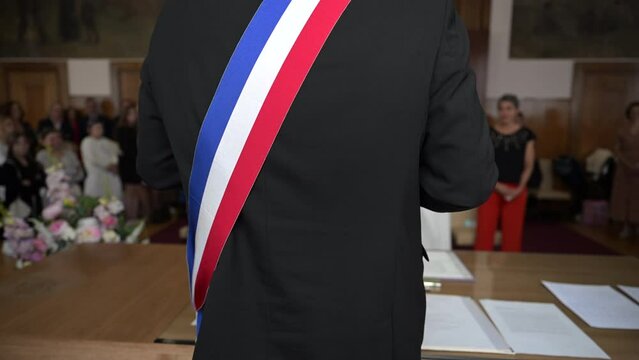 A mayor from behind with a French tricolor scarf making a speech to celebrate a heterosexual marriage in a town hall. Blurred background on the bride and groom. French wedding with two happy lovers.