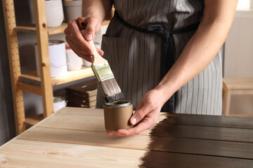 Man dipping brush into can of wood stain at wooden surface indoors, closeup