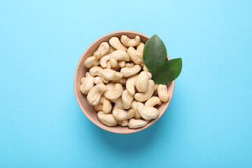 Tasty cashew nuts and green leaves in bowl on light blue background, top view