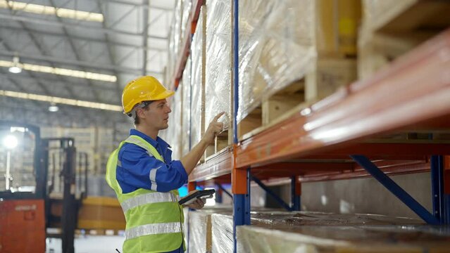 Young caucasian man warehouse workers holding digital tablet checking inventory management packaging boxes. Man staff wearing vest and safety helmet and walking count the box at storehouse