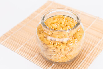 Uncooked Chifferi Rigati Pasta in Glass Jar on Bamboo Mat on White Background. Fat and Unhealthy Food. Classic Dry Macaroni. Italian Culture and Cuisine. Raw Pasta