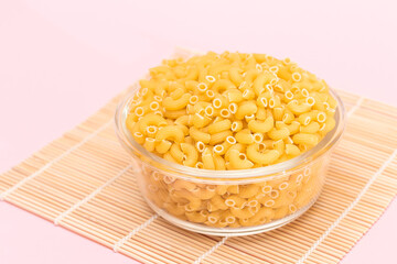 Uncooked Chifferi Rigati Pasta in Glass Jar on Bamboo Mat on Pink Background. Fat and Unhealthy Food. Classic Dry Macaroni. Italian Culture and Cuisine. Raw Pasta