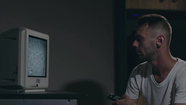 Broken Television or TV Propaganda. Mind Control - Zombified Man in White T-shirt Switches Channels on the Old TV in Dark Room. TV Signal Search. Distortion on the Screen. Medium Close-Up, Film Grain