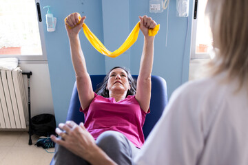 Focused photo of a middle age patient with brown hair while doing exercises with a yellow rubber band and her nurse is in front of her out of focus.