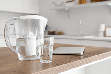 Water filter pitcher, glass and laptop on kitchen table - obrazy, fototapety, plakaty
