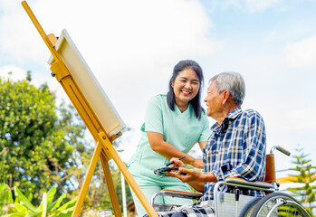 Close up of Asian nurse stand beside senior man sit on wheelchair and draw or paint for relax and...