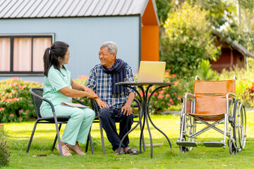 Asian senior man and nurse relax outdoor in the garden with laptop is on the table and wheelchair...