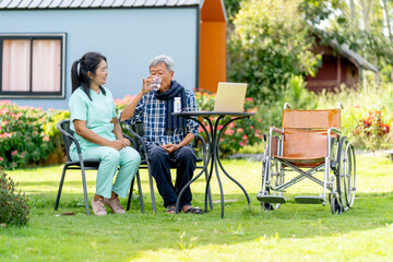 Asian senior man drink water from glass after take the medicine from nurse and they sit outdoor in the garden with wheelchair is set beside.