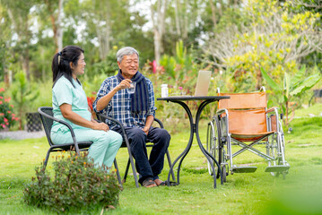 Asian senior man hold a glass of water and talk to nurse after take the medicine from her and they sit outdoor in the garden with wheelchair is set beside.