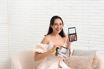 Beautiful young woman with cosmetic bag full of makeup products and eyeshadows palette in room