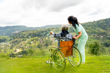 Back of Asian nurse stand and look to the direction of senior man on wheelchair pointing and both stay near cliff with village and mountain in the background.