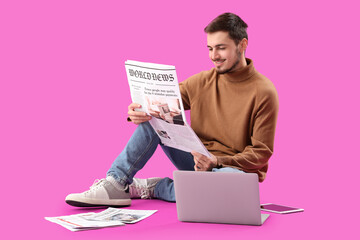 Young man with newspaper and laptop sitting on purple background