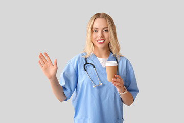 Beautiful female doctor with cup of coffee waving hand on grey background