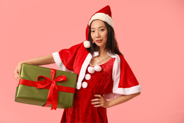 Young Asian woman in Santa costume with Christmas gift box on pink background
