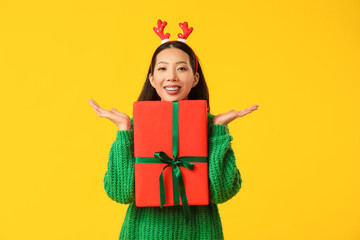 Happy young Asian woman in reindeer horns with Christmas gift box on yellow background
