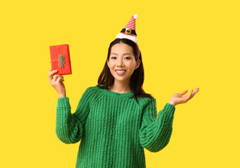 Young Asian woman in Elf hat headband with Christmas gift box on yellow background - obrazy, fototapety, plakaty